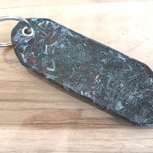 May include: A rectangular keyring with a silver ring. The keyring features a dark, marbled design with shades of green, white, and hints of orange. The surface has a glossy finish.