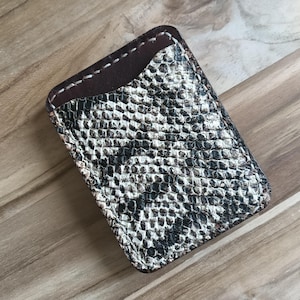 Op de afbeelding: Een rechthoekige kaarthouder met een slangenpatroon in zwart, wit en grijs. Het bovenste gedeelte is effen donkerbruin en de randen zijn gestikt met witte draad. De houder is gemaakt van leer.
