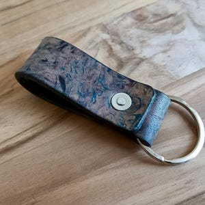 Könnte beinhalten: Ein Leder-Schlüsselanhänger mit einem marmorierten Muster in Braun-, Blau- und Grüntönen. Der Schlüsselanhänger hat einen silberfarbenen Metallring und einen Druckknopfverschluss. Der Lederriemen ist etwa 10 cm lang.