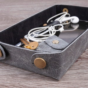 Puede incluir: Una bandeja de cuero gris con un forro de fieltro negro. La bandeja está llena de varios artículos, incluyendo llaves, un teléfono y auriculares.