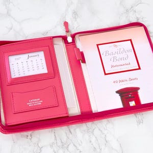 May include: A pink leather zippered folder with a calendar, a card holder, and a notepad. The notepad has the text "Basildon Bond Watermarked" and "40 White Sheets".