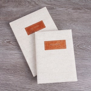 May include: Two white linen journals with brown leather covers and the text "MY DIARY" embossed on the covers. The journals are on a wooden surface.