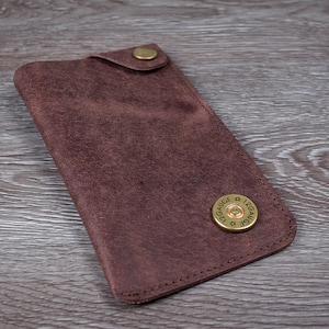 May include: A brown leather wallet with a 12 gauge shotgun shell button. The wallet is lying on a wooden surface.