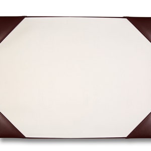 May include: A rectangular desk blotter with a cream writing surface and dark brown leather corners. The corners are triangular and stitched to the edges. The blotter is designed to protect a desk surface.
