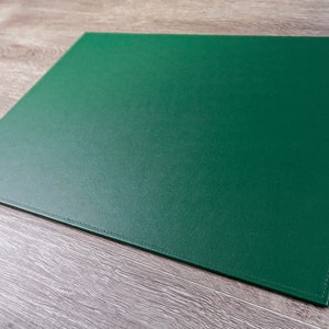 May include: A green cutting mat with stitched edges. The mat is lying on a light brown wooden surface.