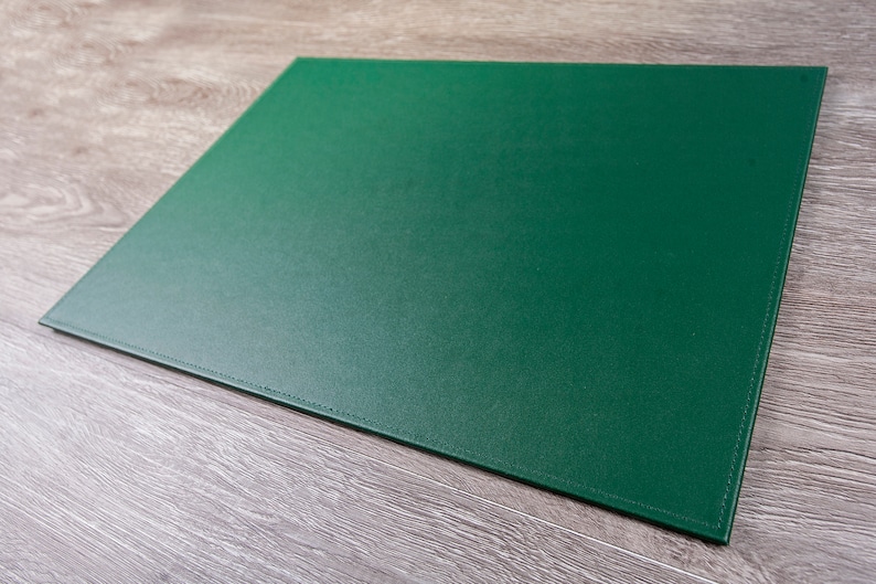May include: A green cutting mat with stitched edges. The mat is lying on a light brown wooden surface.