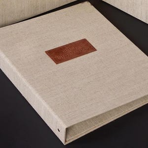 May include: A beige linen-covered binder with a brown leather patch that reads "GUEST INFORMATION." The rectangular binder has metal rings on the spine. The design is simple and elegant, suitable for various uses.