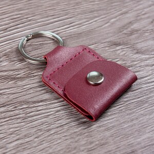 May include: A burgundy leather keychain with a small pocket and a silver metal ring. The keychain has a silver metal button.