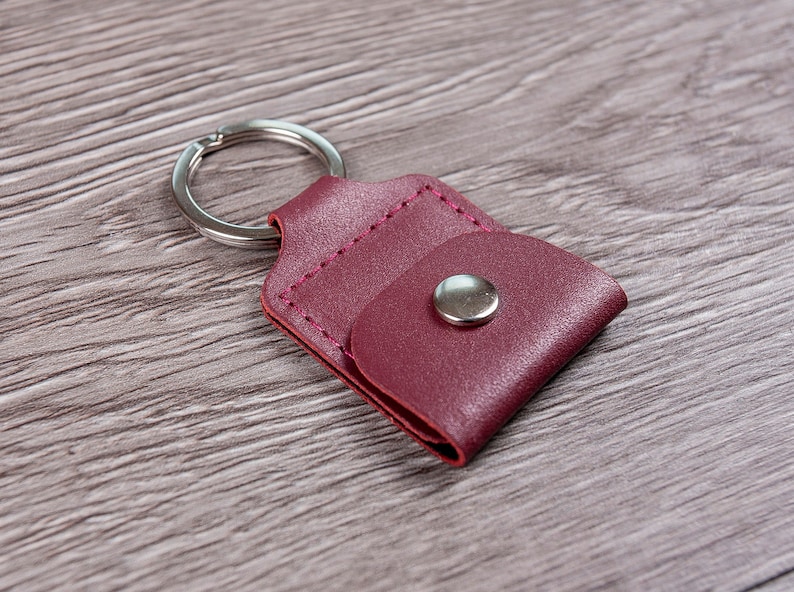 May include: A burgundy leather keychain with a small pocket and a silver metal ring. The keychain has a silver metal button.