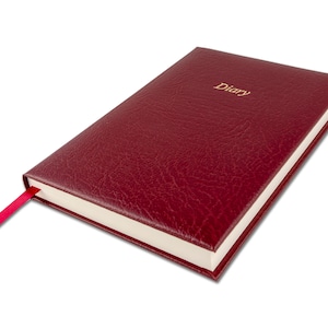 Könnte beinhalten: Ein burgunderfarbenes, ledergebundenes Tagebuch mit einer goldenen Aufschrift "Diary". Das Tagebuch hat ein rotes Lesezeichenband.