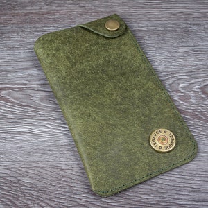 May include: Olive green leather phone case with a brass button closure. The case features a brass 12-gauge shotgun shell casing accent. The case is rectangular and has a distressed finish, suggesting a rugged aesthetic.