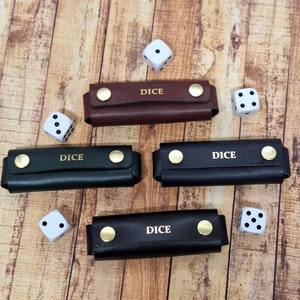 May include: Three black leather dice holders with gold accents and the word "DICE" embossed on them. One brown leather dice holder with gold accents and the word "DICE" embossed on it. Six white dice with black dots are scattered around the holders.