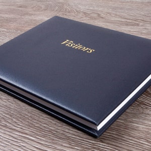 May include: A navy blue guest book with the word "Visitors" in gold lettering. The book has a square shape and a textured cover. The pages have white edges.