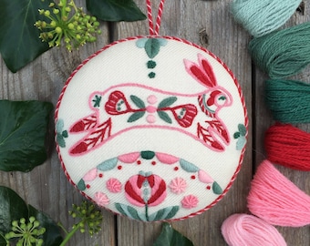 Könnte beinhalten: Ein weißer Stoffschmuck mit einem gestickten Hasenmotiv in Rosa und Rot. Der Schmuck ist von grünen Blättern und rosa, roten und grünen Stickgarnen umgeben.