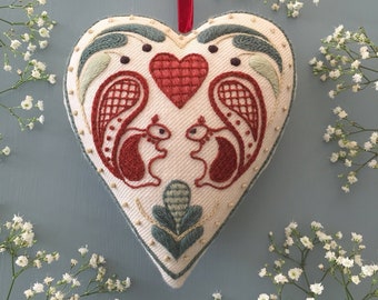 May include: A white heart-shaped cushion with red and green embroidery. The embroidery depicts two squirrels facing each other with a red heart in the centre.