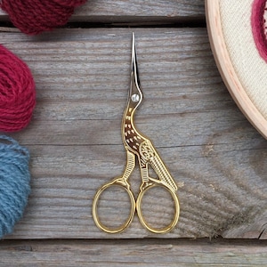Puede incluir: Tijeras de bordado doradas con un diseño de pájaro. Las tijeras están sobre una superficie de madera con ovillos de hilo rojo, azul y blanco.