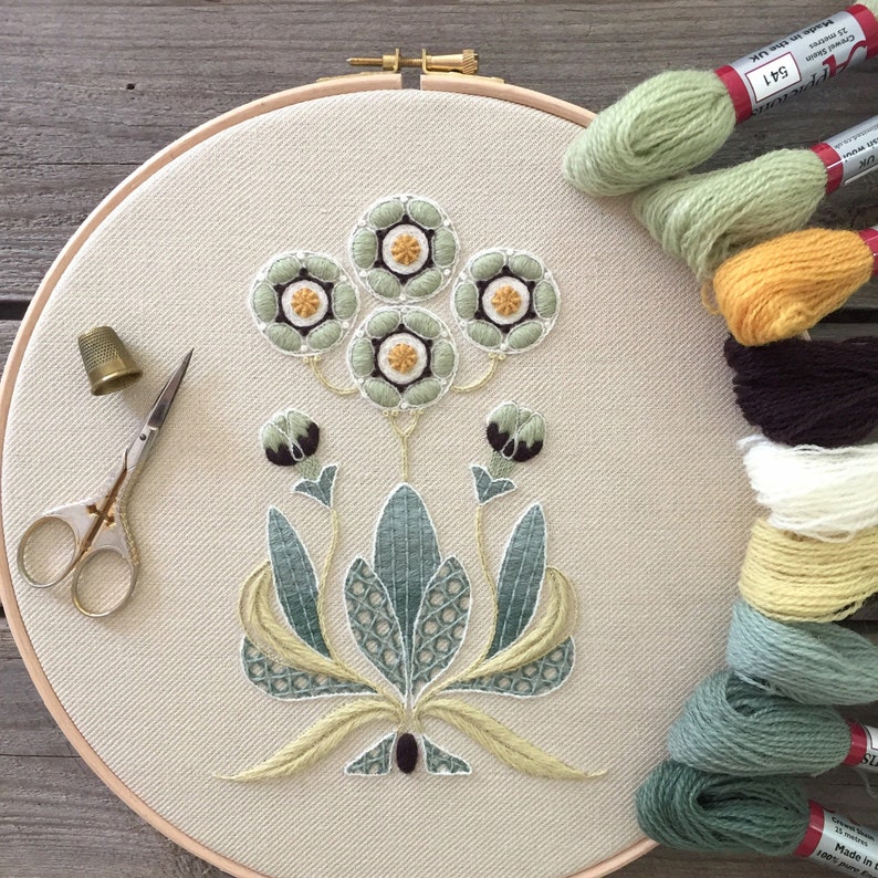 May include: A close-up of a hand-stitched embroidery hoop with a floral design. The design features five green and white flowers with yellow centers, surrounded by green leaves. The hoop is on a wooden surface with several skeins of embroidery floss in various colors.