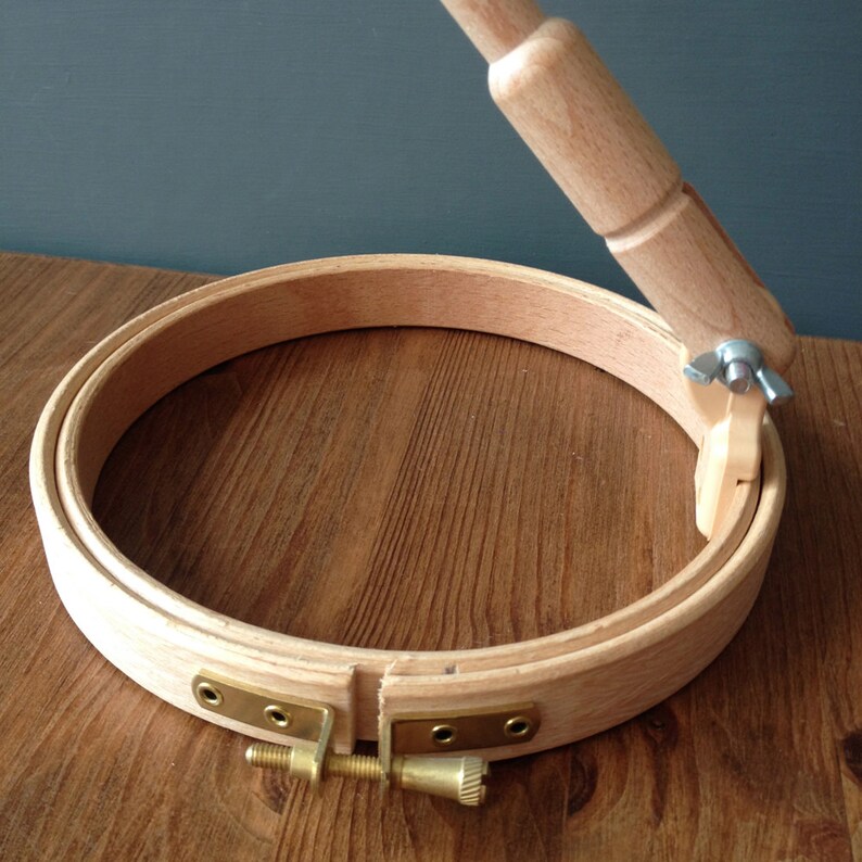 May include: A wooden embroidery hoop with a tightening mechanism. The hoop is made of natural wood and has a brass screw and nut.
