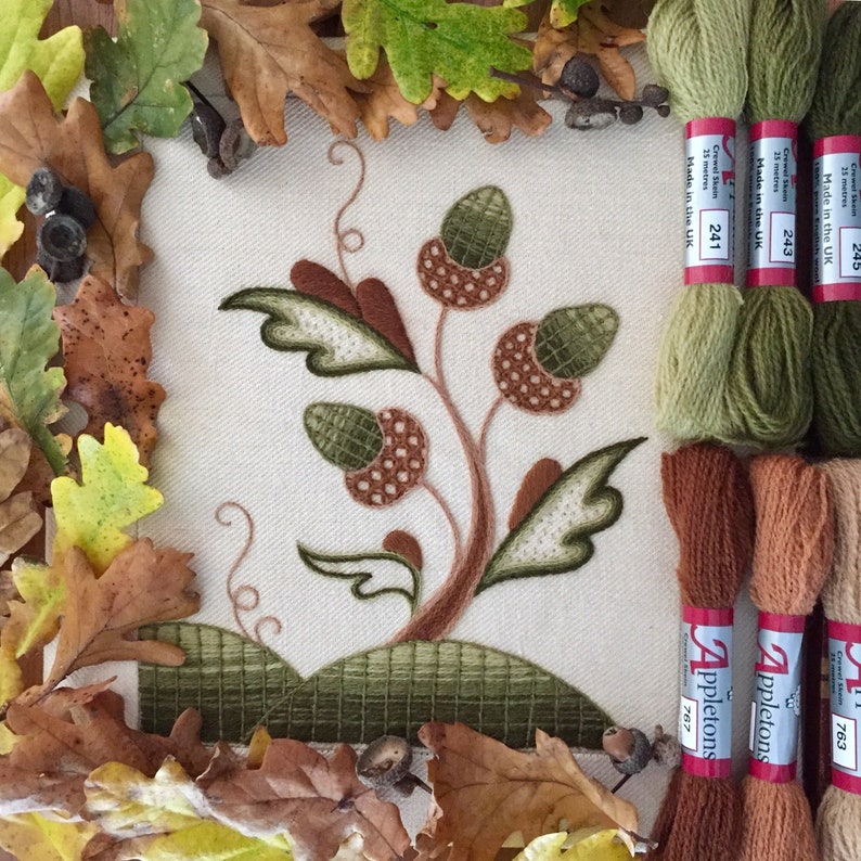 May include: A close-up of a needlepoint embroidery project depicting a stylized tree with acorns. The embroidery is framed by a border of brown and green leaves. The image also shows three spools of green embroidery floss and three spools of brown embroidery floss, labeled "Appletons" and with numbers "763", "767", and "241".