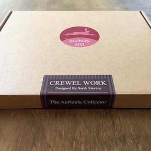 May include: A brown cardboard box with a red and white logo that says "Melbury Hill" and a brown label that says "CREWEL WORK Designed By Sarah Stevens The Auricula Collector"