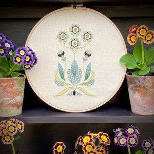 May include: A white embroidery hoop with a floral design featuring green and yellow leaves and white flowers with yellow centers. The hoop is on a shelf with potted plants with purple flowers.