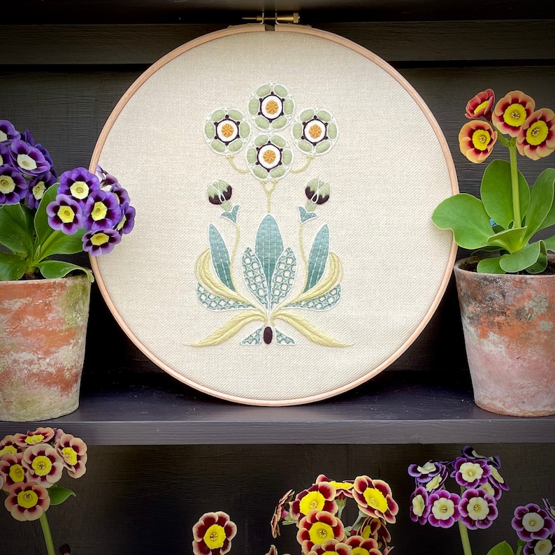 May include: A white embroidery hoop with a floral design featuring green and yellow leaves and white flowers with yellow centers. The hoop is on a shelf with potted plants with purple flowers.