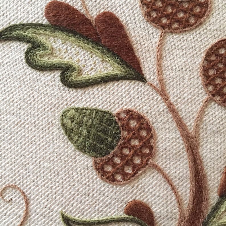 May include: Close-up of a brown and green embroidered design on a white fabric. The design features a leafy stem with acorns and a brown and green leaf.