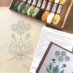 May include: A close-up of a needlepoint embroidery kit featuring a floral design with the text "The Auricula Collector". The kit includes a piece of linen fabric, embroidery floss in various colors, and a needle. The kit is packaged in a brown box with a white label.