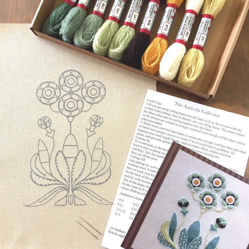 May include: A close-up of a needlepoint embroidery kit featuring a floral design with the text "The Auricula Collector". The kit includes a piece of linen fabric, embroidery floss in various colors, and a needle. The kit is packaged in a brown box with a white label.
