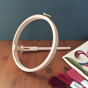 Puede incluir: Un bastidor de bordado de madera con cierre de latón y brazo de madera ajustable. El bastidor está sobre una superficie de madera, con madejas de hilo de bordar rojo y rosa y una tela bordada.