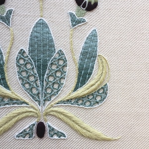 May include: Close-up of a hand-stitched embroidery design featuring a stylized floral motif with green and yellow thread on a cream-colored fabric.