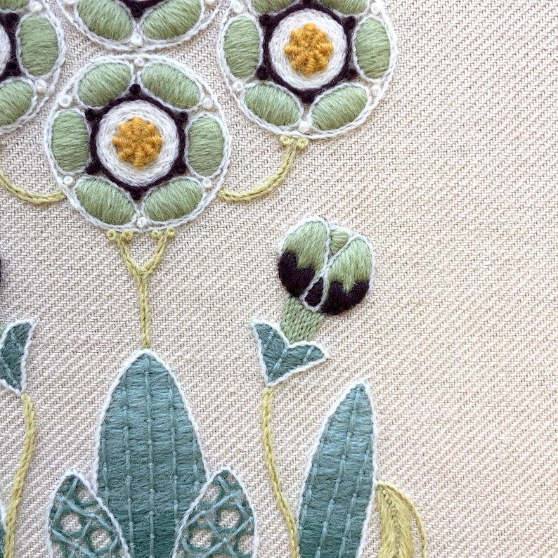 May include: Close-up of an embroidered design featuring green, white, and brown flowers with yellow centers. The flowers are on a beige fabric background.