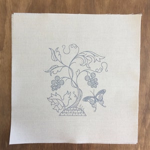 May include: A white linen fabric with a gray embroidered design of a vine with grapes and a butterfly. The vine is stylized with a basket at the base.