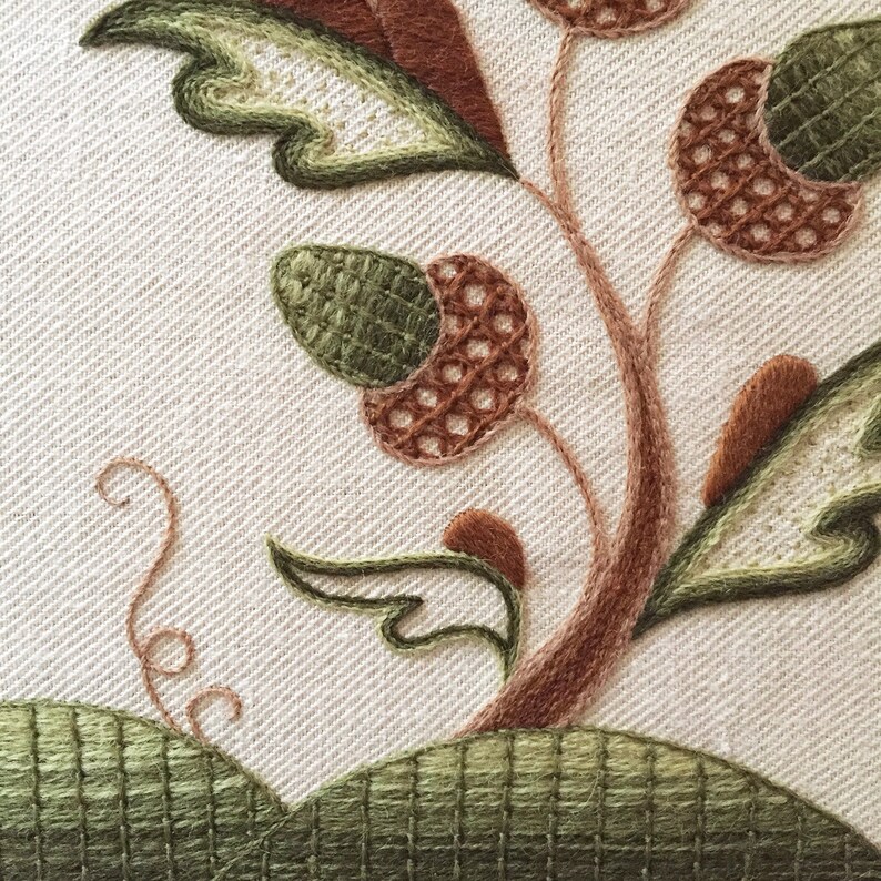 May include: Close-up of a brown and green embroidered design featuring a stylized tree with acorns and leaves. The embroidery is stitched on a white linen fabric.