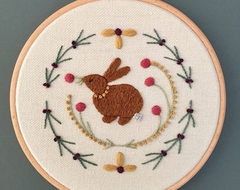 May include: Embroidered hoop art featuring a brown bunny with a gold necklace, surrounded by green leaves, pink flowers, and gold beads.