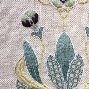 May include: Close-up of a hand-stitched embroidery design featuring a green and brown flower with green leaves on a beige fabric background.