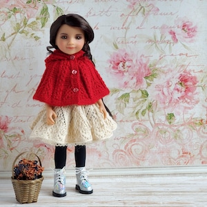 May include: A doll wearing a red knitted cape with a hood and a cream-colored lace skirt. The doll has black tights and silver boots. A small wicker basket with dried flowers sits nearby. The background features a floral wallpaper.