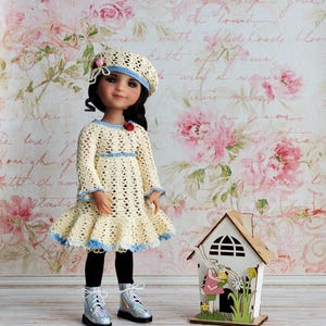 May include: A doll wearing a cream-colored crochet dress with blue trim, a matching beret, and silver shoes. The doll is standing in front of a floral wallpaper and next to a small wooden house.