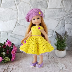 Op de afbeelding: Een pop gekleed in een gele gebreide jurk, een paarse baret en bijpassende paarse schoenen. De jurk heeft een gedetailleerd ajourpatroon en een kleine paarse hartvormige versiering. De baret is versierd met een gele hart.