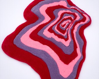 May include: A pink, purple, and red abstract rug with a wavy, organic shape. The rug has a layered, concentric design.