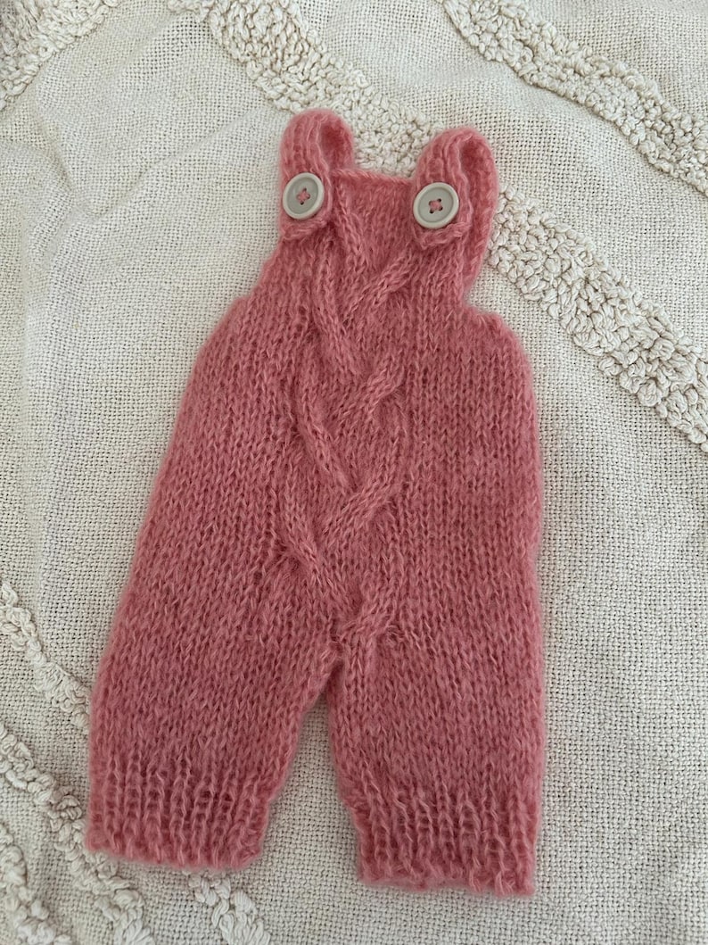 Pu&ograve; includere: Una tutina in maglia color rosa polvere con un motivo a trecce centrale, due bottoni circolari di colore chiaro sulle spalline e polsini a coste sulle aperture delle gambe. Il capo &egrave; realizzato in un filato morbido e soffice.