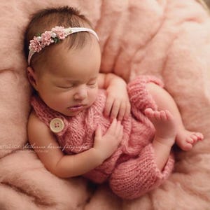 Pu&ograve; includere: Un neonato che dorme indossa una tutina in maglia rosa polvere con un bottone chiaro sulla bretella e una fascia abbinata con piccoli fiori di stoffa rosa e perle bianche, adagiato su un tessuto rosa morbido e testurizzato.