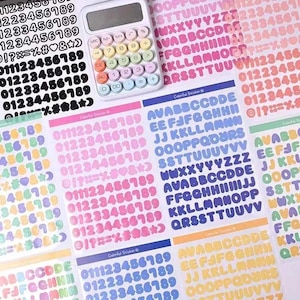 Op de afbeelding: Een witte rekenmachine met kleurrijke knoppen ligt op vellen kleurrijke stickers. De stickers bevatten cijfers, letters en symbolen in verschillende kleuren, waaronder roze, blauw en groen. De stickers zijn voor knutselen.