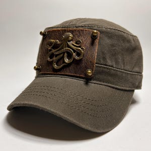 May include: Olive green military-style cap with a brown leather patch featuring a bronze octopus design. The patch is secured with four brass rivets. The cap has a short brim and a structured crown.