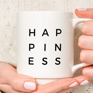 May include: White ceramic mug with black text that reads "Happiness" in a unique font style. The mug is being held by a person's hands.