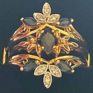 May include: A gold-toned ring with a floral design. The ring features dark blue marquise-cut gemstones and small, round, clear gemstones. The ring has an intricate, layered design with filigree details.