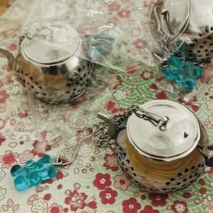 May include: Three silver-colored metal tea infusers shaped like miniature teapots. Each infuser features a perforated body, a small lid, and a silver chain attached to a translucent blue gummy bear-shaped charm. Two are inside clear plastic bags.
