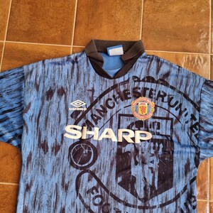Puede incluir: Una camiseta de f&uacute;tbol azul y negra con el logotipo del Manchester United y la palabra "SHARP" impresa en la parte delantera. La camiseta est&aacute; hecha por Umbro.