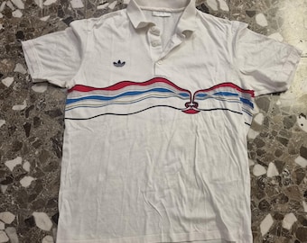May include: White polo shirt with a colorful horizontal design across the chest. The design includes red, blue, and yellow stripes. The shirt has a collar and short sleeves, and the Adidas logo is on the left chest.