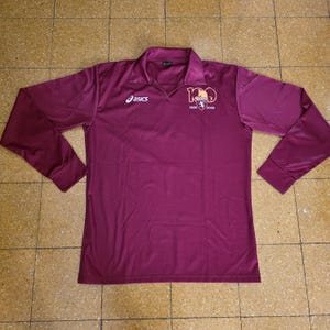 May include: A long-sleeved, burgundy athletic shirt with a collared neckline. The Asics logo is on the left chest, and a crest with the text "1906-2006" is on the right chest. The shirt is made of a smooth, likely synthetic material.